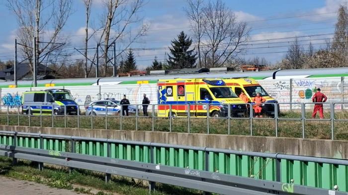 Zespół ratunkowy z ambulansami i radiowozem zabezpiecza miejsce zdarzenia obok torów kolejowych.