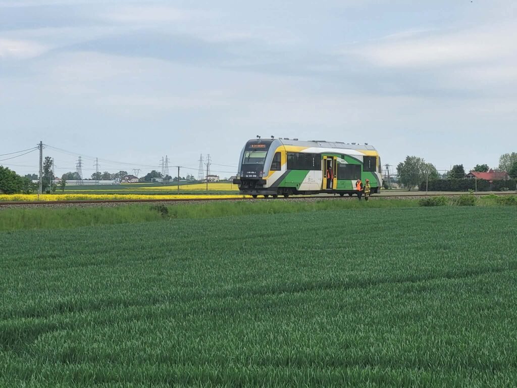 Pociąg regionalny zatrzymany na torach wśród zielonych pól, a obok dwóch pracowników w odzieży roboczej.