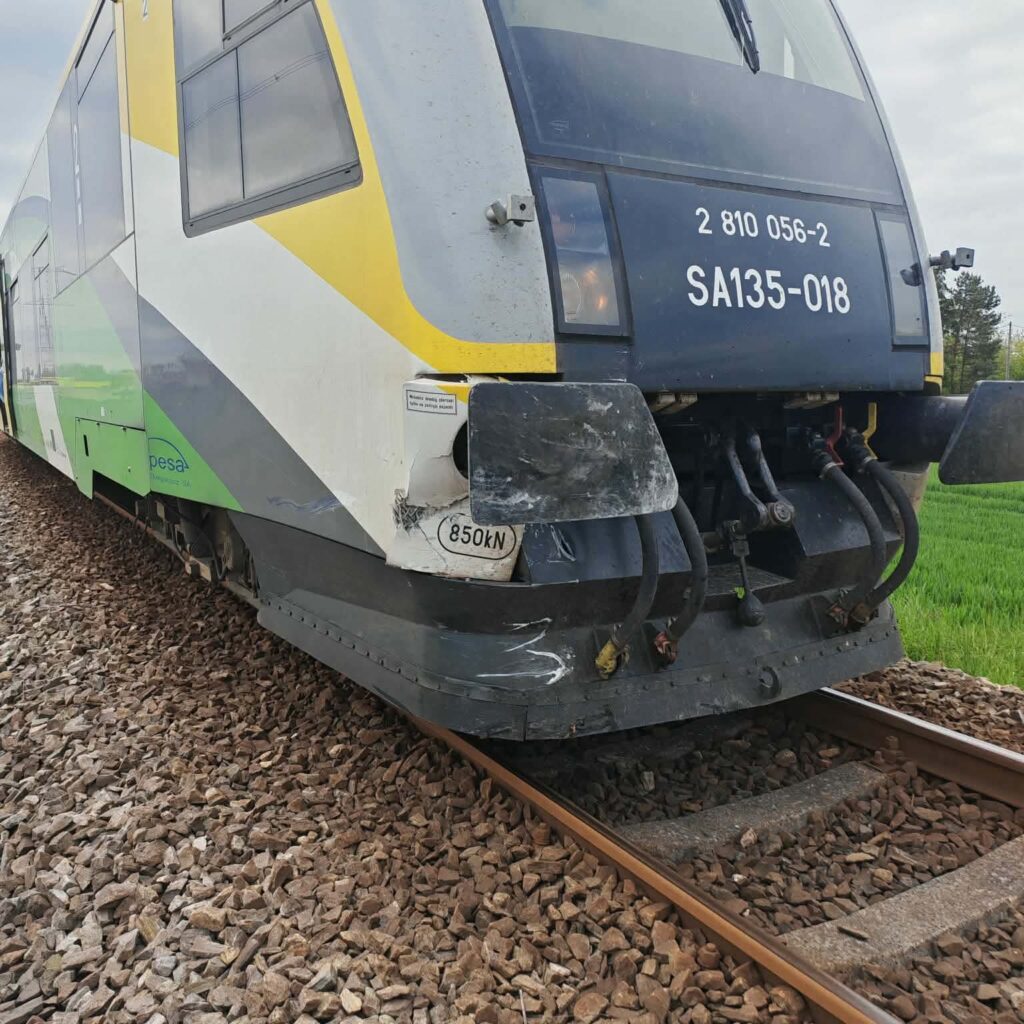 Pociąg spalinowy z uszkodzoną przednią częścią stojący na torach otoczony zielonymi polami.