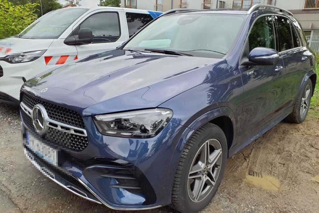 Niebieski SUV marki Mercedes zaparkowany na żwirowym podjeździe, obok białego samochodu dostawczego.