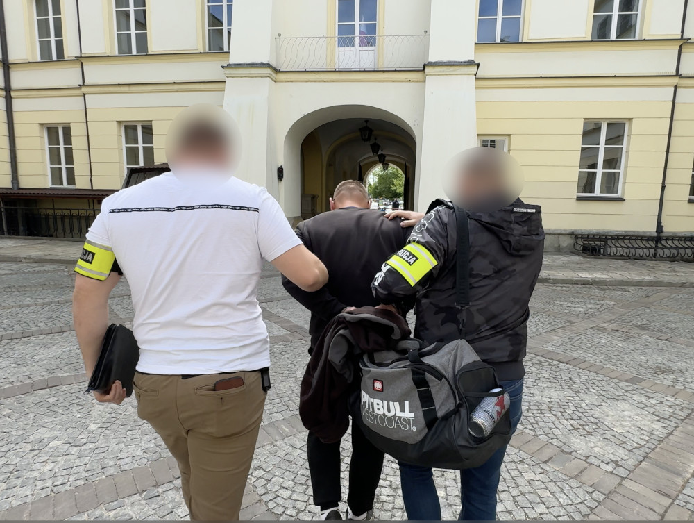 Trzech mężczyzn w odzieży z oznaczeniami "POLICJA" prowadzi jednego mężczyznę przez brukowaną przestrzeń w architektonicznym otoczeniu.