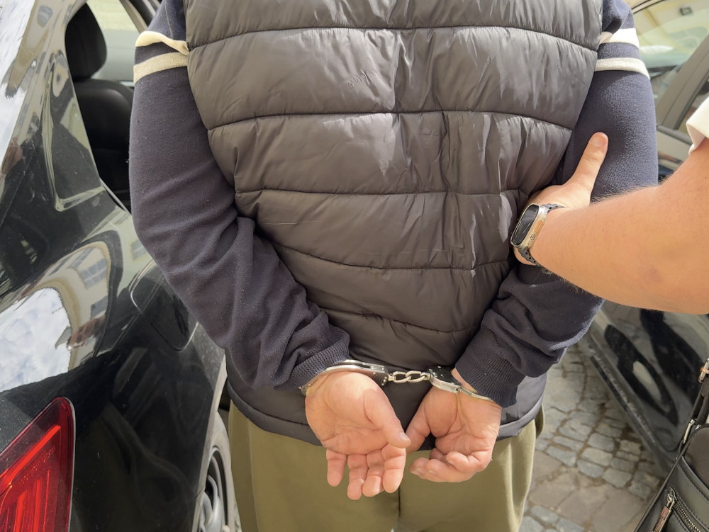 Mężczyzna w kajdankach stoi przy czarnym samochodzie, z rękami z tyłu, podczas interwencji policji.