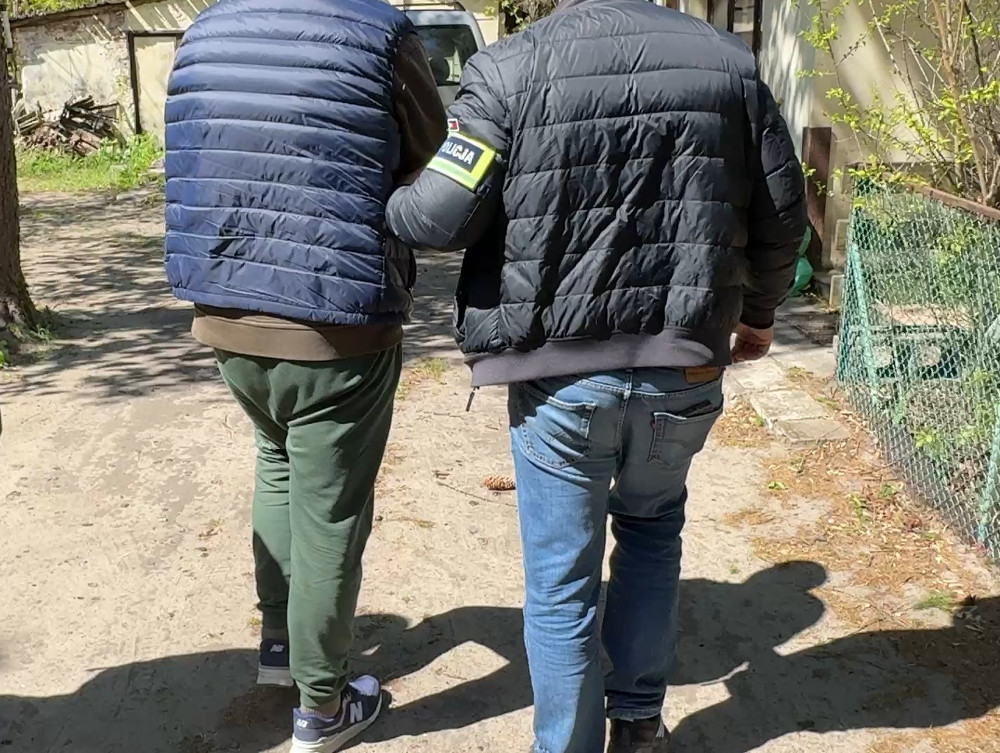 Dwaj mężczyźni w puchowych kurtkach spacerują po ścieżce w otoczeniu zieleni i budynków.