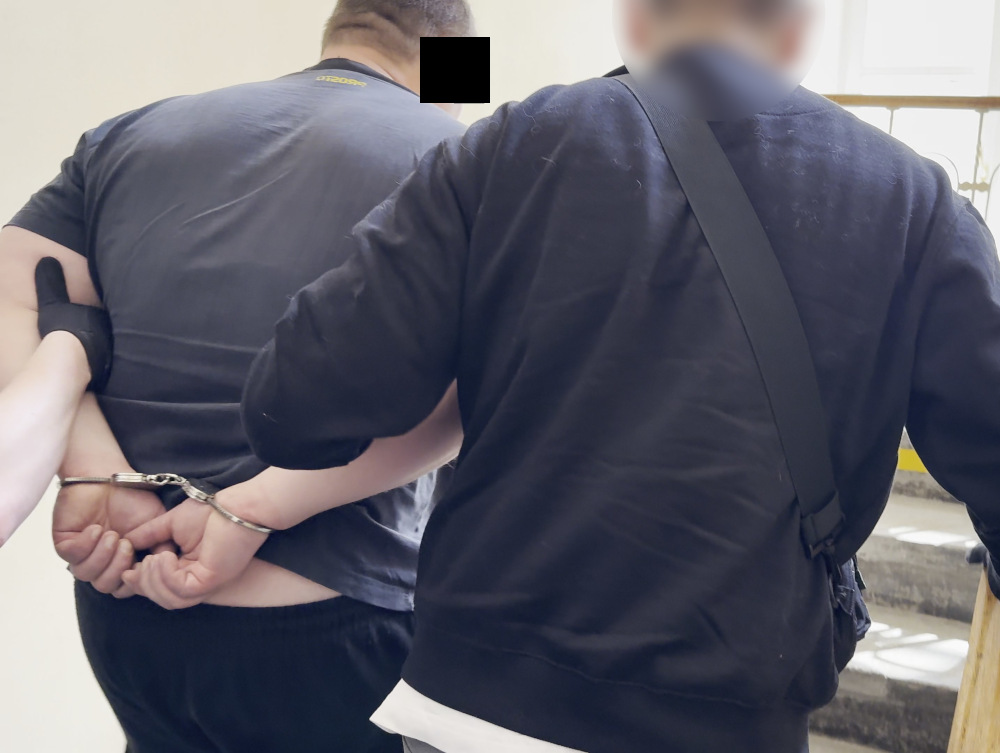 Mężczyzna w kajdankach prowadzony przez funkcjonariusza na schodach budynku.