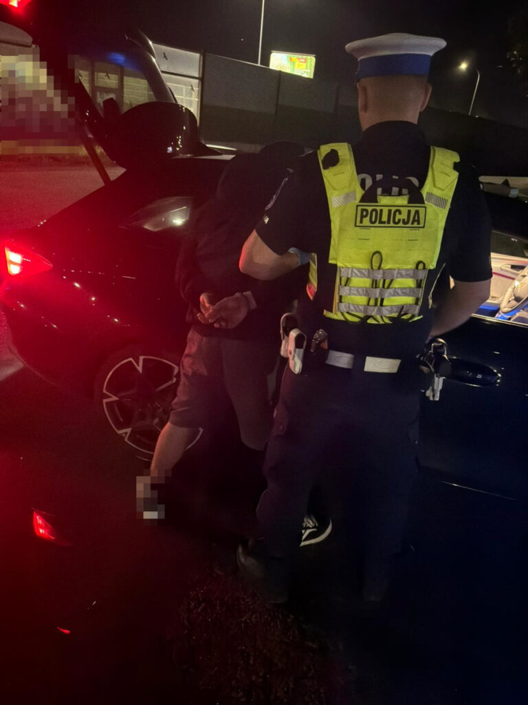 Policjant w kamizelce odblaskowej zatrzymuje mężczyznę w czarnej bluzie nocą przy otwartym bagażniku samochodu.