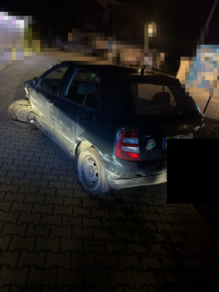 Zniszczony samochód osobowy stoi na wyłożonej kostką drodze, w nocy, z jednym uszkodzonym kołem.