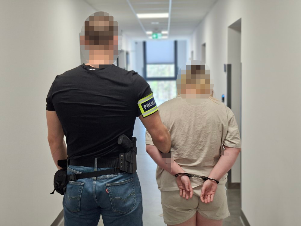 Policjant w mundurze prowadzi aresztowaną osobę z rękami skrępowanymi z tyłu w korytarzu budynku.