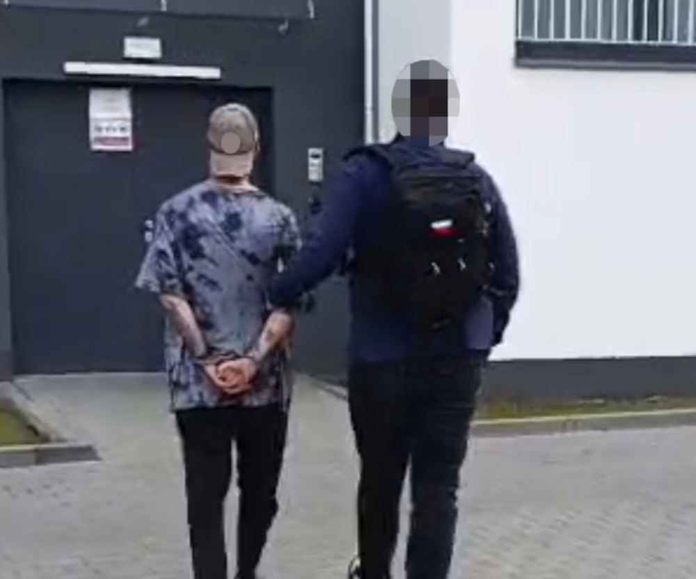 Mężczyzna z rękami związanymi z tyłu idzie w towarzystwie funkcjonariusza w kierunku budynku.