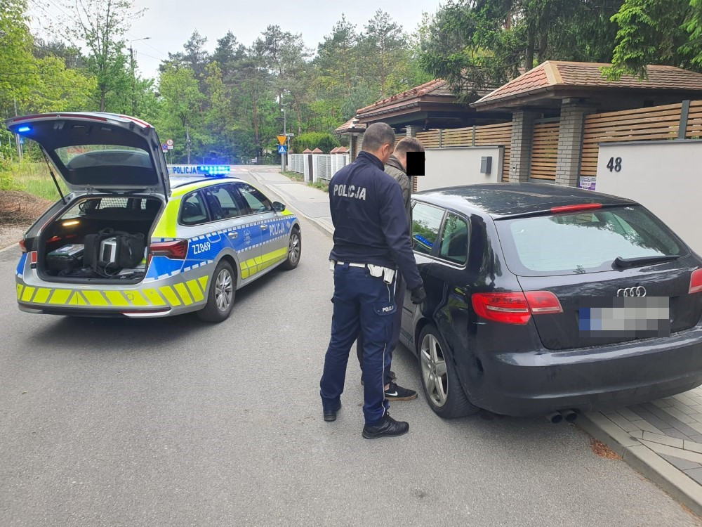 Policjant kontrolujący czarne auto na ulicy, obok radiowozu z włączonymi sygnałami świetlnymi.
