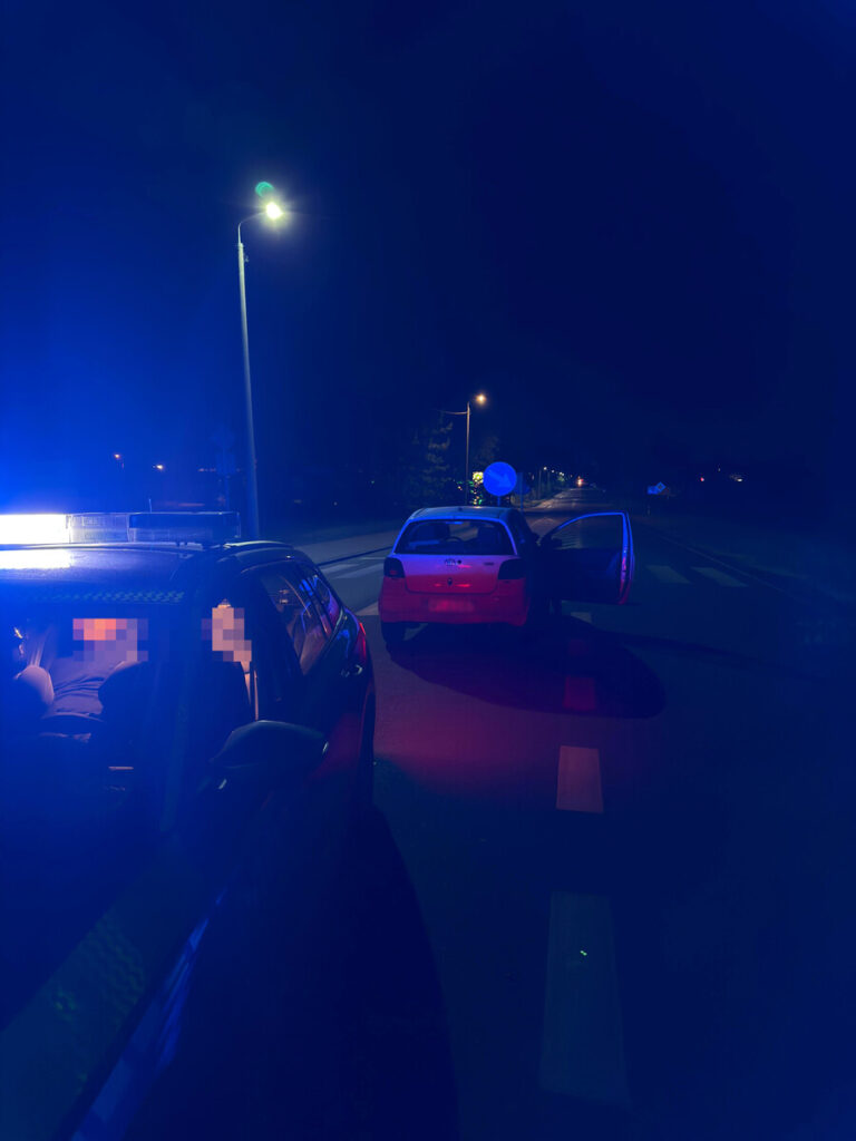 Policyjny radiowóz z włączonymi sygnałami świetlnymi stoi na drodze nocą, obok otwartego samochodu osobowego.