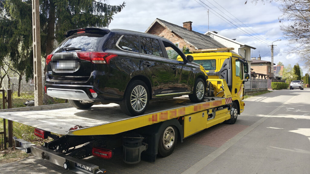 Czarny SUV załadowany na żółtą lawetę na ulicy w otoczeniu domów jednorodzinnych.