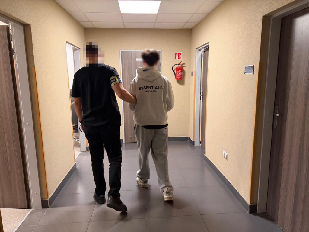 Mężczyzna w czarnym ubraniu prowadzi młodego mężczyznę w szarym dresie korytarzem w budynku.