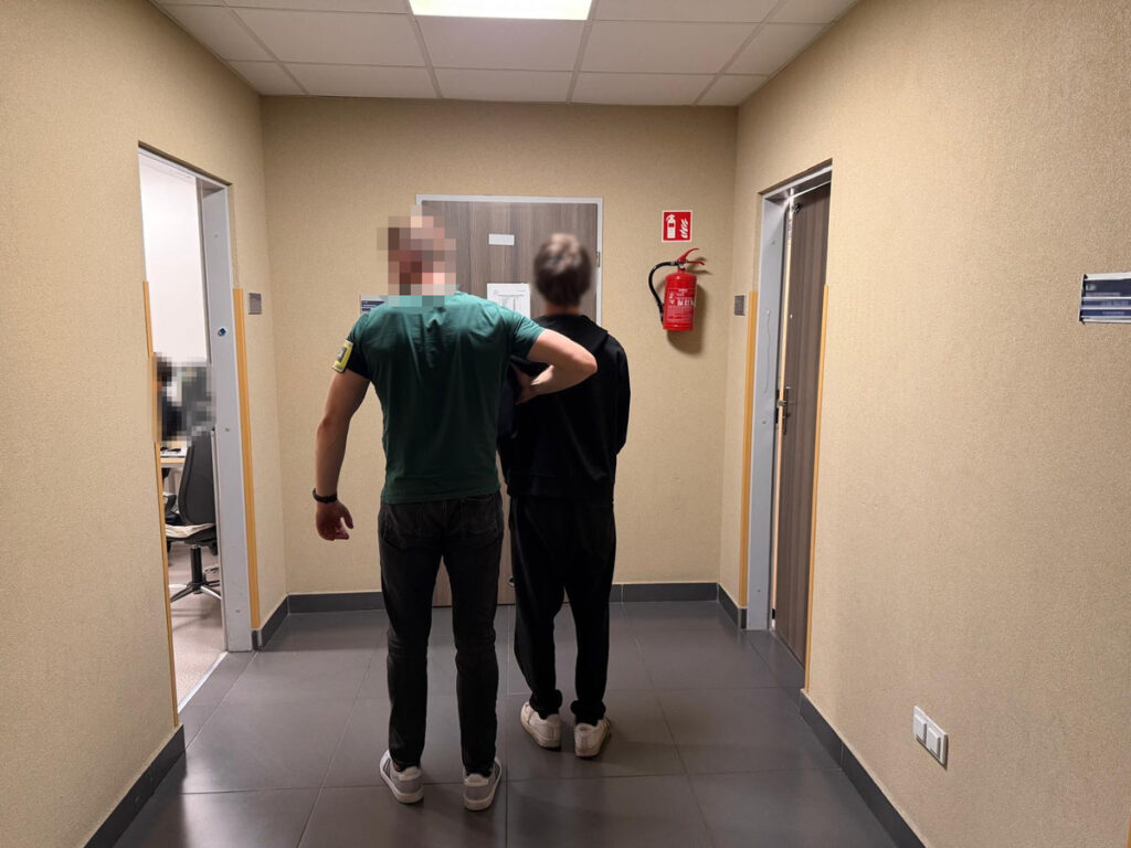 Mężczyzna w zielonej koszulce prowadzi drugiego mężczyznę w czarnej odzieży korytarzem w budynku biurowym.