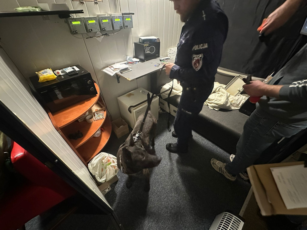Policjant z psem służbowym w pomieszczeniu, w tle biurko z urządzeniami elektronicznymi i półka z przedmiotami.