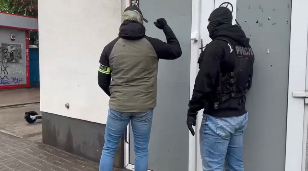 Mężczyzna w kamizelce taktycznej i maską stoi obok innego mężczyzny w garniturze, puka do drzwi budynku.