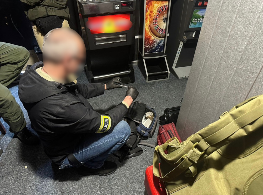 Funkcjonariusz w czarnym płaszczu z emblematem "POLICJA" przeszukuje torbę w lokalu z automatami do gier.