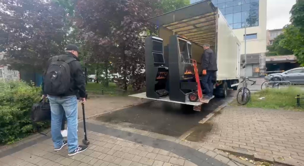 Mężczyzna z torbą obserwuje dwóch pracowników rozładowujących automaty do gier z ciężarówki na chodniku.