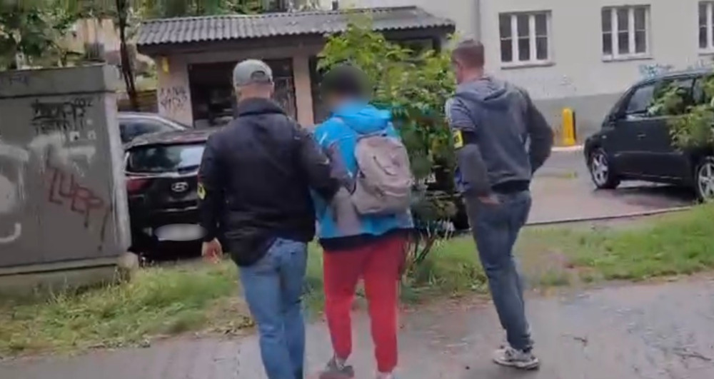Dwaj mężczyźni w odzieży roboczej prowadzą osobę z plecakiem w kierunku budynku w miejskim otoczeniu.