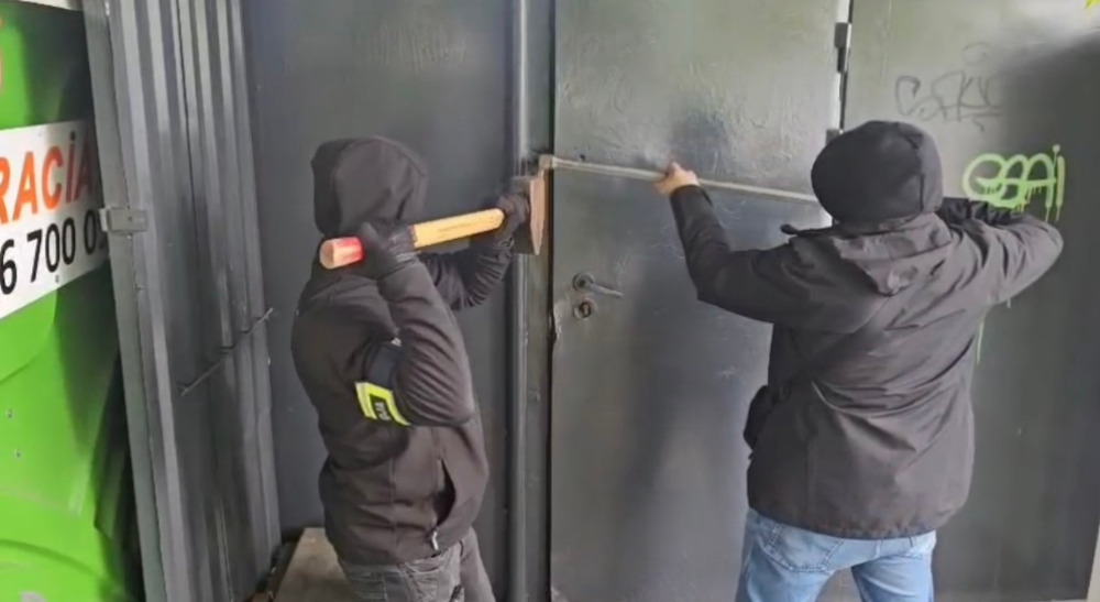 Dwaj mężczyźni w kapturach próbują otworzyć metalowe drzwi przy użyciu łomu i młota.