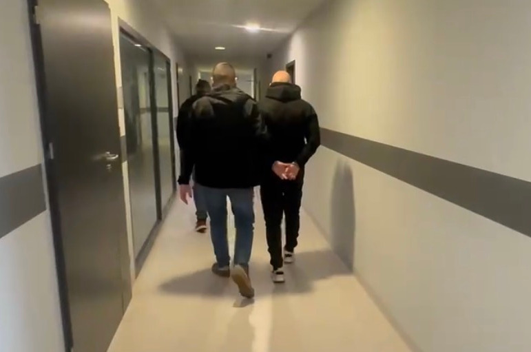 Mężczyźni idą korytarzem, jeden z nich z rękami z tyłu, w otoczeniu jasnych ścian i zamkniętych drzwi.