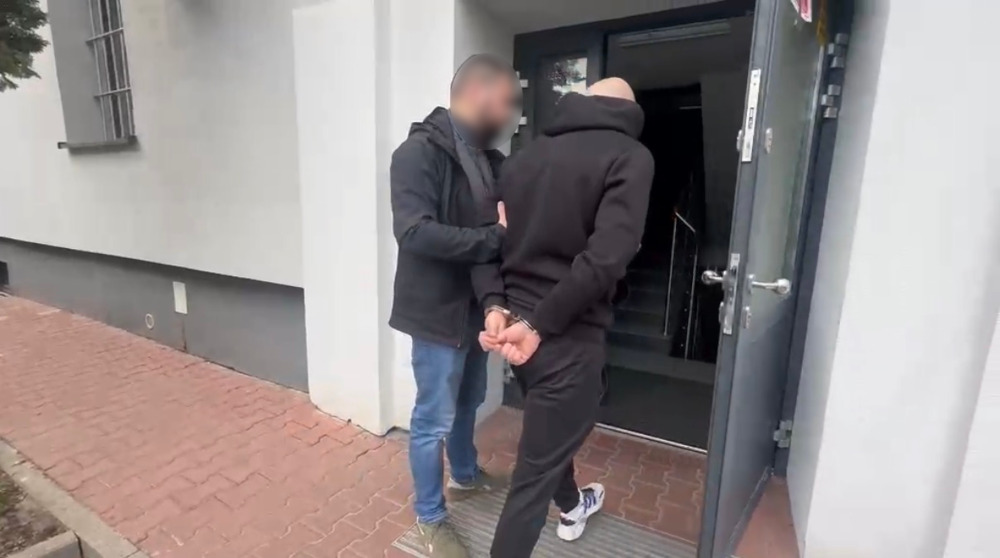 Mężczyzna w czarnym dresie prowadzony przez funkcjonariusza policji przy drzwiach budynku.