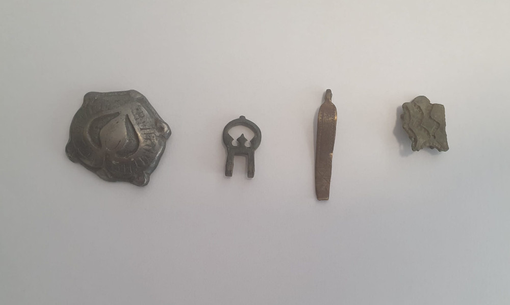 Cztery różne przedmioty metalowe leżące na białym tle: ozdobny medalion, mały symbol, prosty wisiorek i fragment z wizerunkiem ptaków.