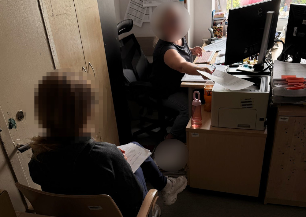 Kobieta siedząca przy biurku przegląda dokumenty, podczas gdy druga kobieta w biurze wskazuje na papiery.
