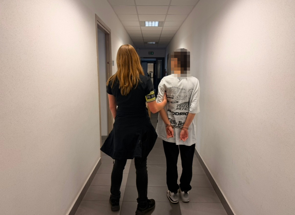 Funkcjonariusz policji prowadzi zatrzymaną osobę w korytarzu budynku, obie mają związane ręce.