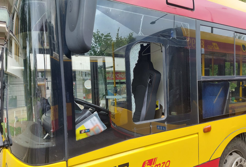 Zniszczona szyba w drzwiach autobusu miejskiego z widoczną uszkodzoną kierownicą wewnątrz pojazdu.