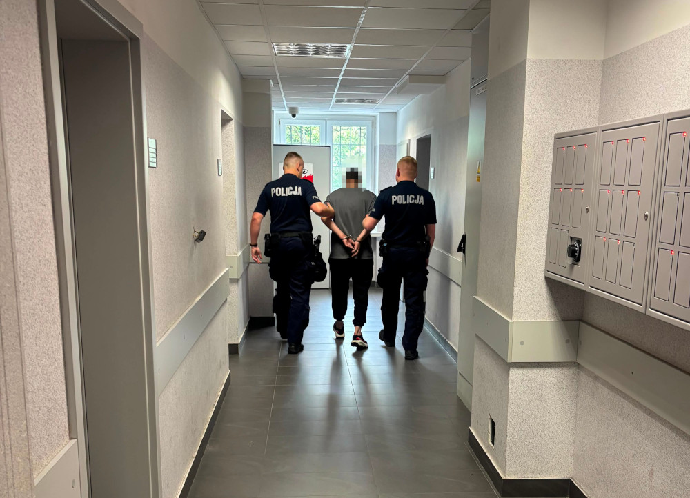 Dwaj policjanci prowadzą mężczyznę w kajdankach przez korytarz w budynku.