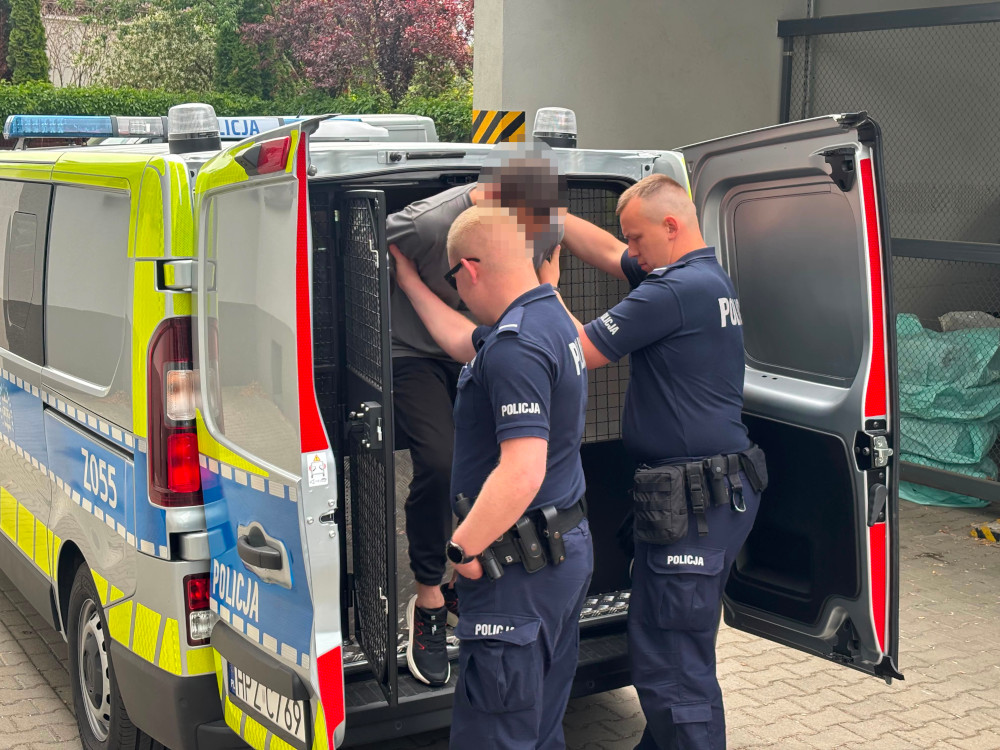 Dwaj policjanci pomagają mężczyźnie wsiąść do furgonetki policyjnej na parkingu.