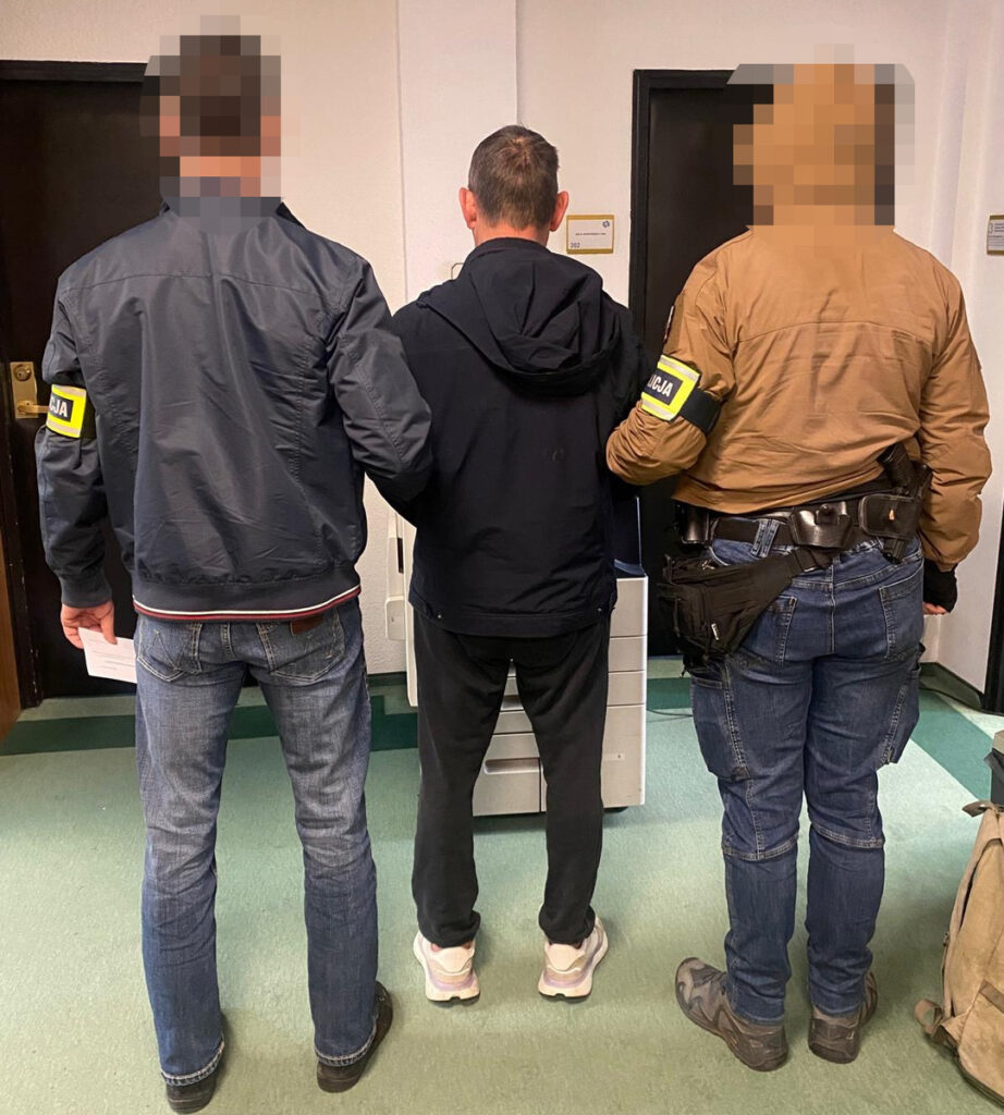 Trzech mężczyzn w cywilnych ubraniach prowadzi aresztowanego mężczyznę w czarnym dresie w korytarzu budynku.
