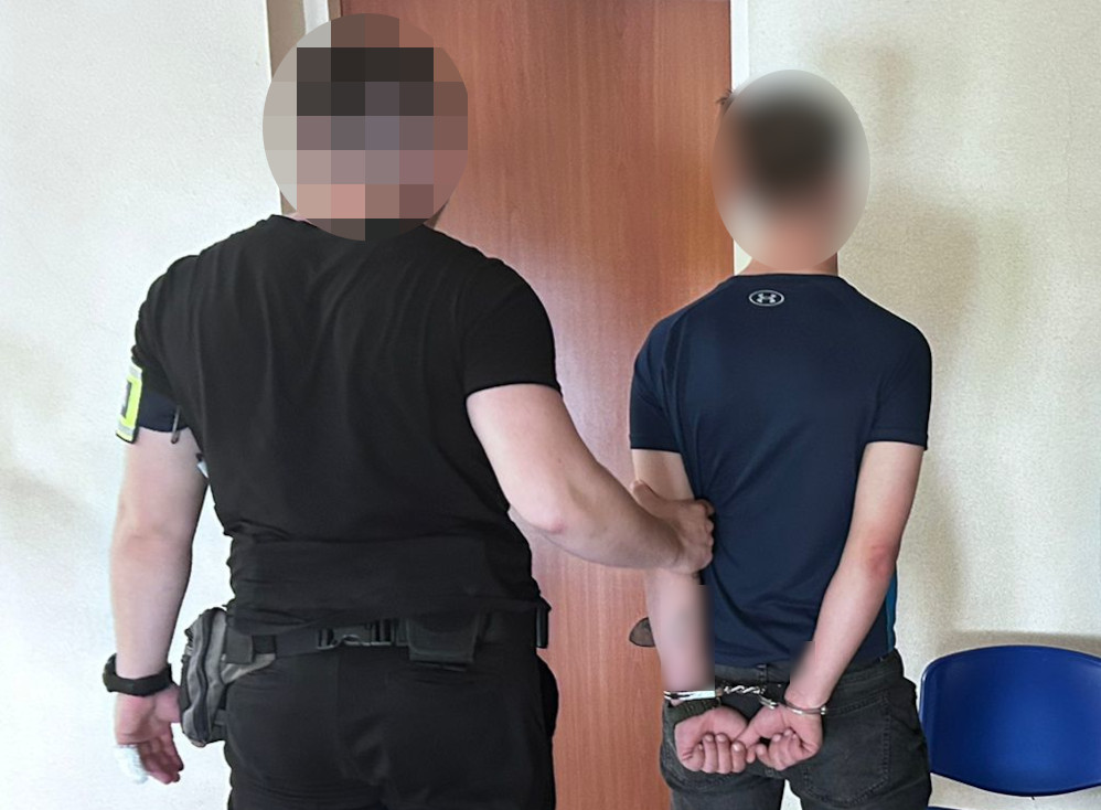 Funkcjonariusz policji zatrzymuje młodego mężczyznę z rękami w kajdankach w pomieszczeniu z zamkniętymi drzwiami.
