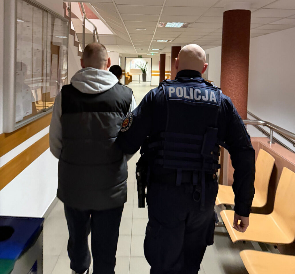 Mężczyzna w szarej bluzie prowadzony przez policjanta w mundurze korytarzem budynku.