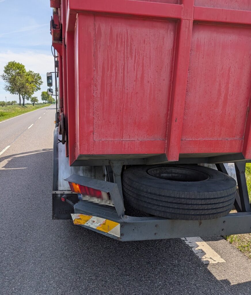 Czerwony ciężarówka z uszkodzoną tylną częścią stoi na drodze, obok widoczne jest pole i drzewa.