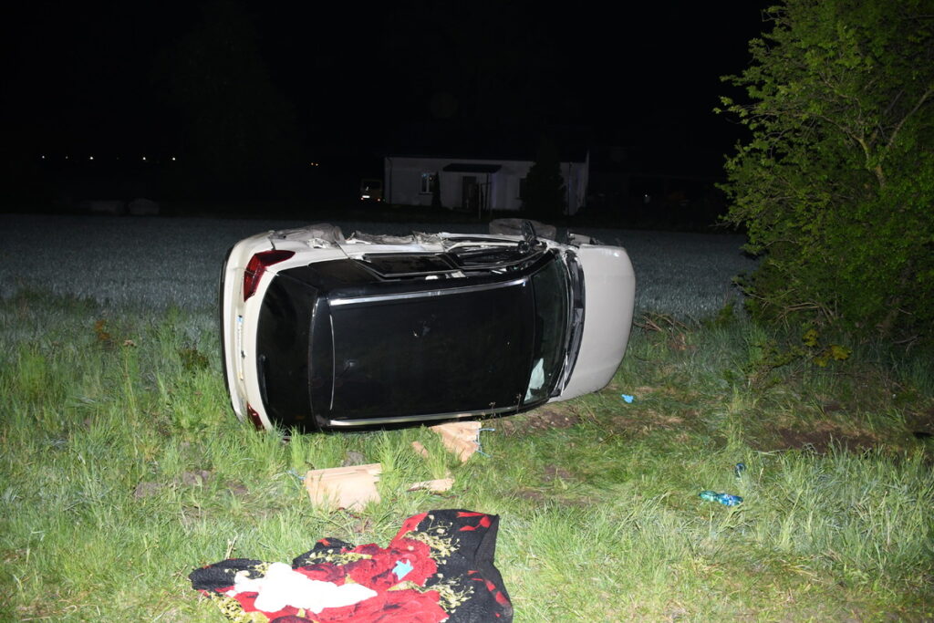 Samochód leżący na dachu w zielonym terenie, obok leżą fragmenty ubrania oraz inne przedmioty. Nocna sceneria.