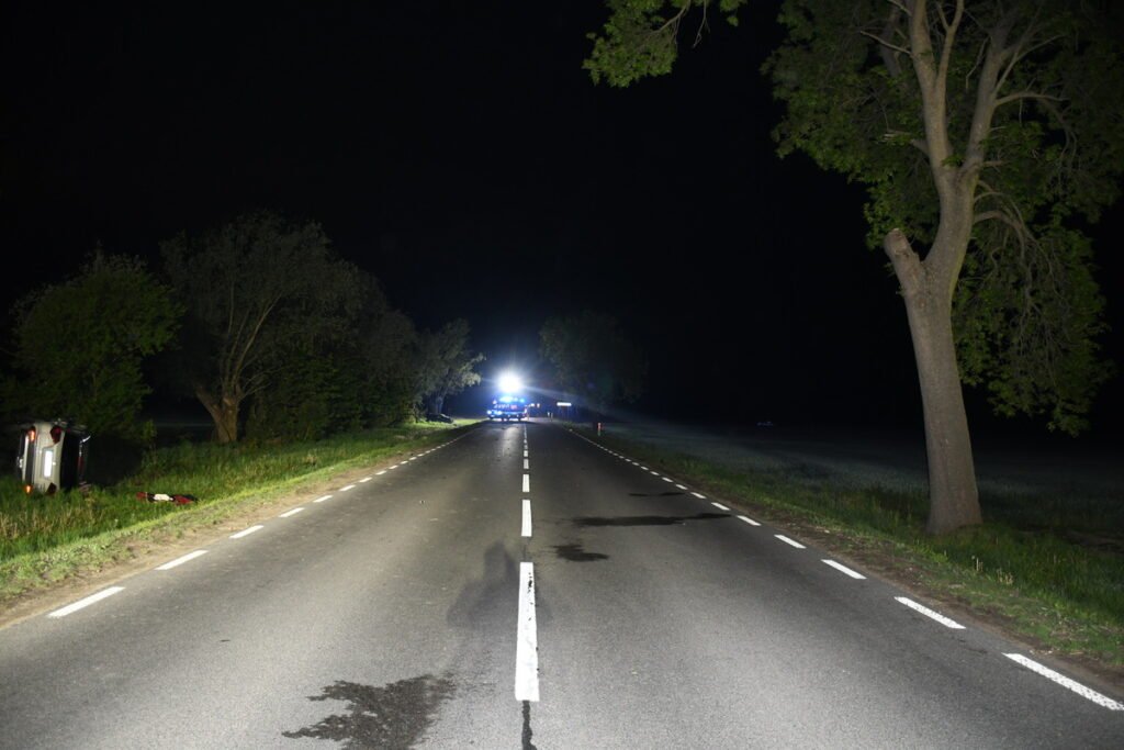 Na drodze nocą leży przewrócone auto, obok widać plamy oleju; w tle świecą niebieskie światła radiowozu.