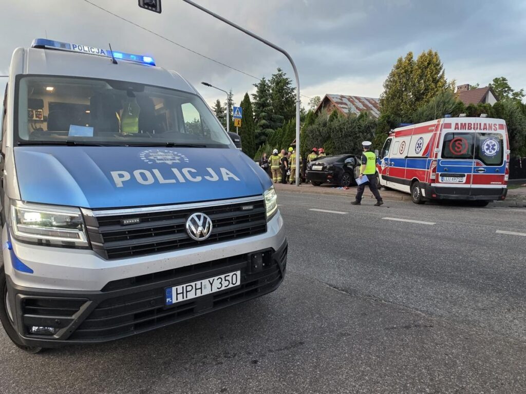 Policyjny van zatrzymany na ulicy, obok karetki pogotowia, gdzie strażacy udzielają pomocy poszkodowanemu w wypadku.