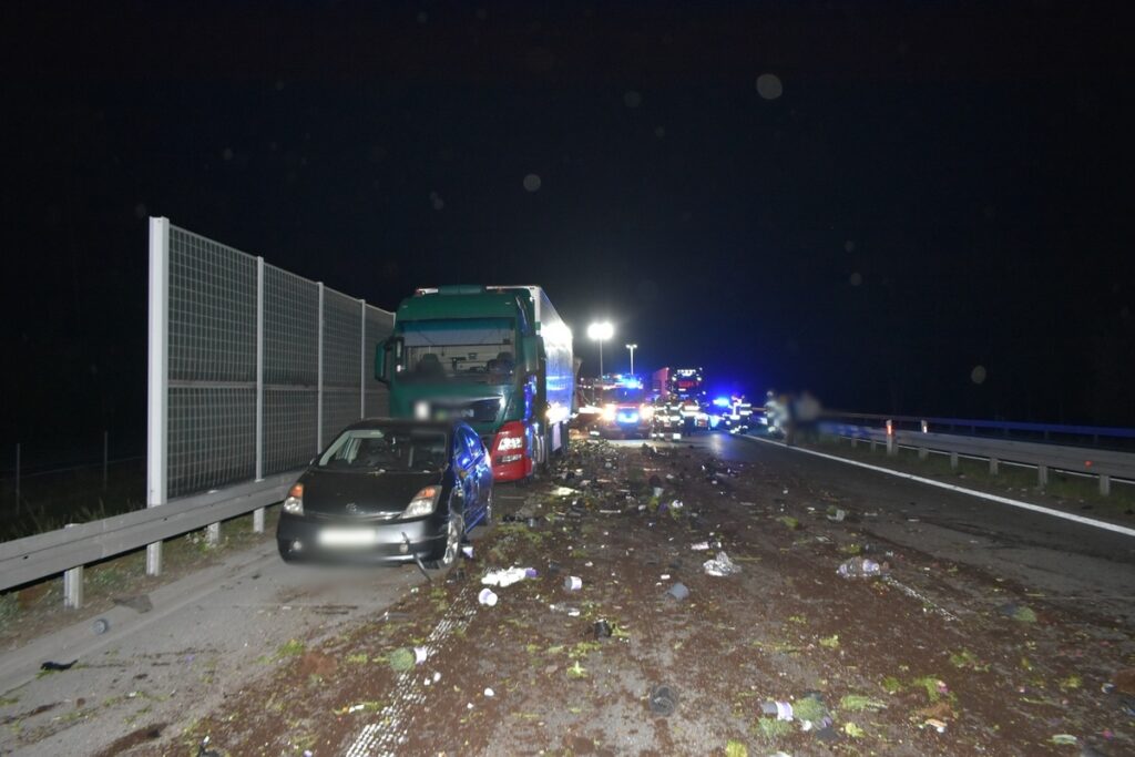 Na drodze nocą stoi uszkodzony czarny samochód obok dużego niebieskiego ciężarowego, w tle widać pojazdy straży pożarnej.