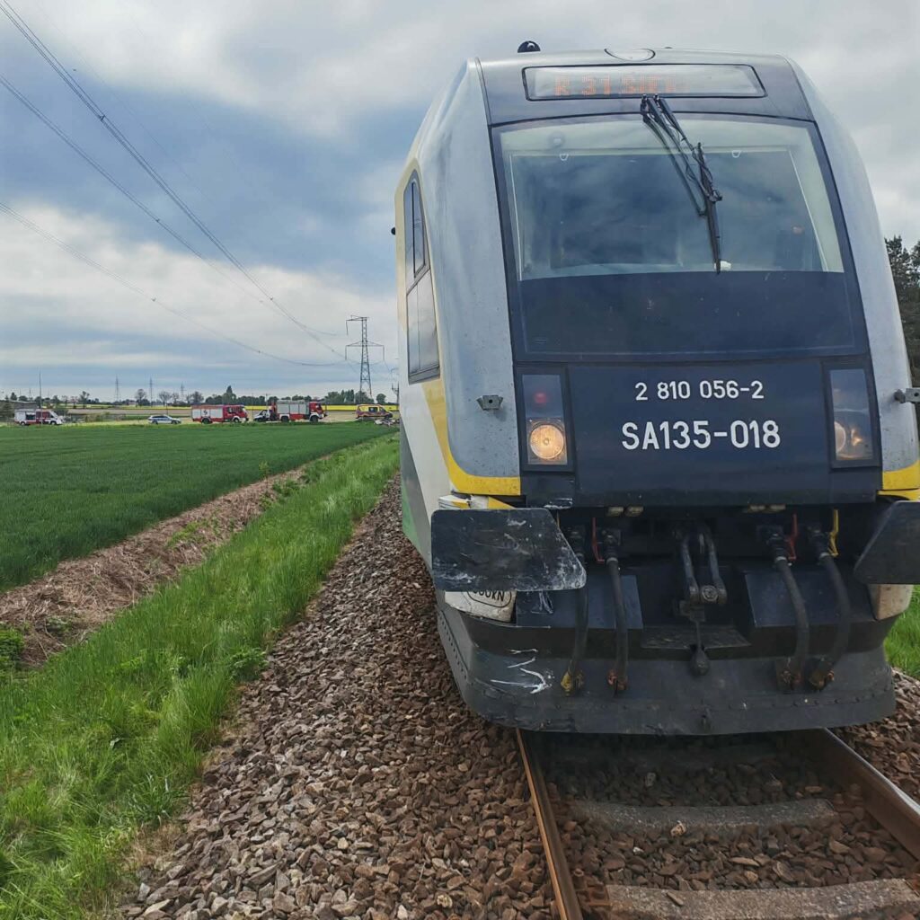 Pociąg osobowy na torach, w tle widoczne pojazdy straży pożarnej i zielone pola.