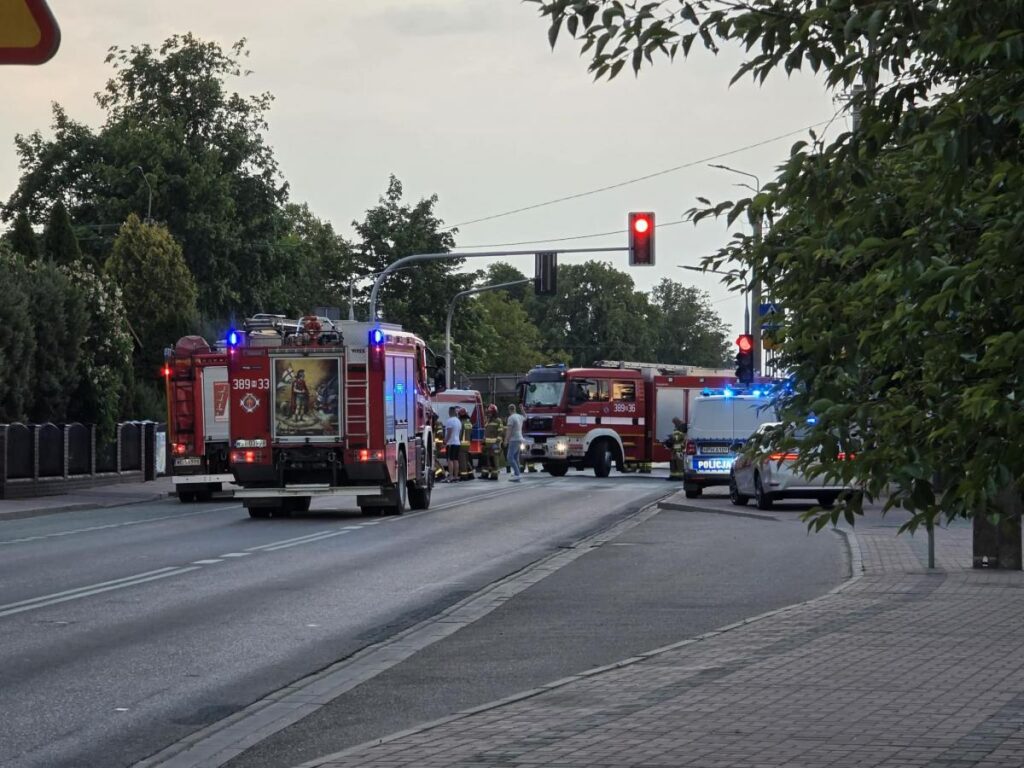 Na ulicy parkują dwa wozy strażackie z włączonymi sygnalizacjami, obok nich zespół ratowników.