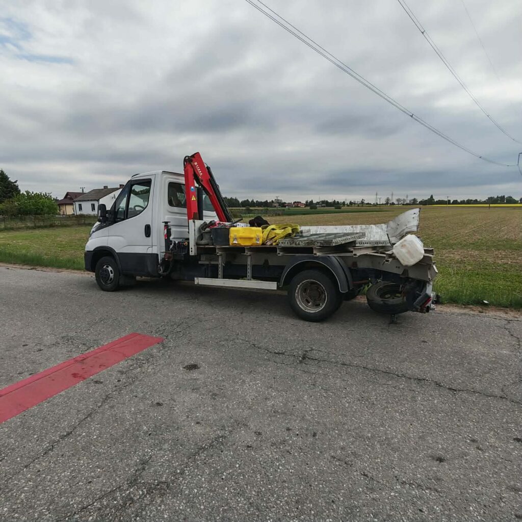 Biały samochód dostawczy z podnośnikiem hydraulicznym zaparkowany na poboczu drogi, obok pola.