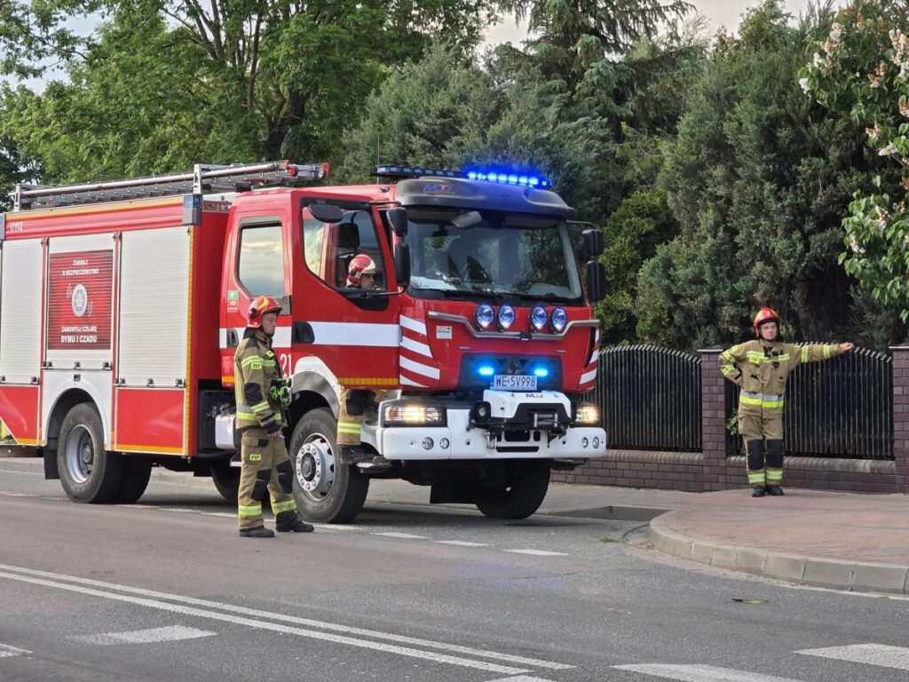 Strażacy w mundurach kierują ruchem obok wozu strażackiego z włączonymi sygnałami świetlnymi.