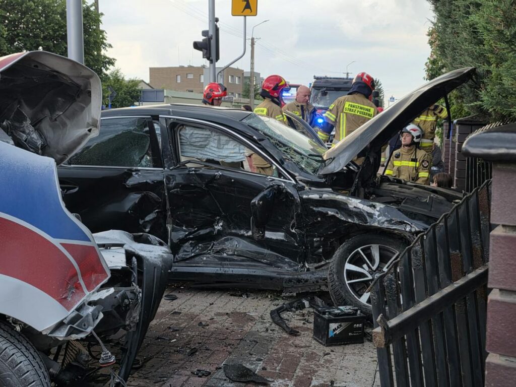 Czarna limuzyna zniszczona w wypadku, obok strażacy w mundurach i policjanci na miejscu zdarzenia.