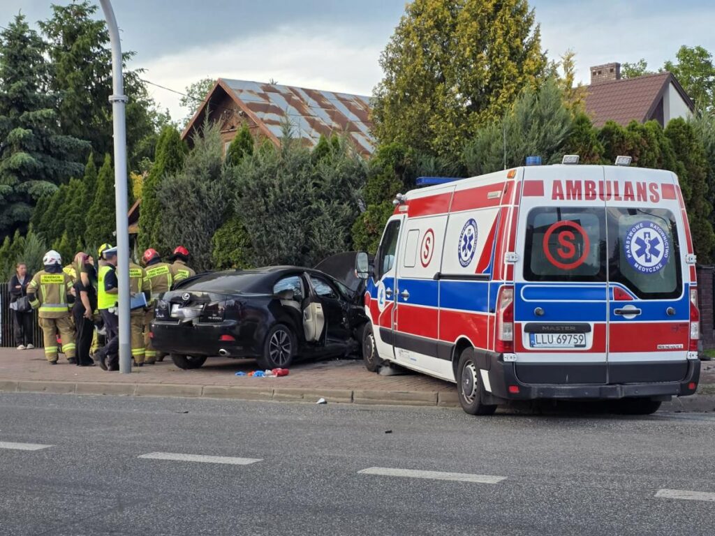 Grupa ratowników medycznych i strażaków pracuje przy rozbitym samochodzie na chodniku, obok stoi ambulans.