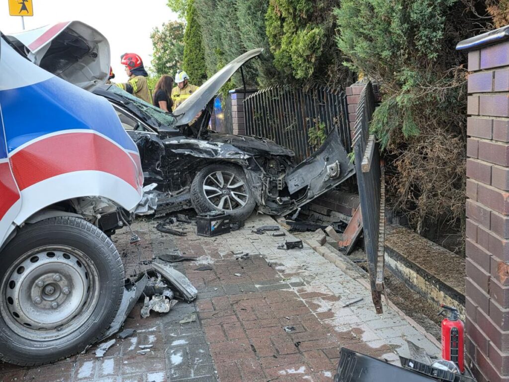 Wypadek drogowy, uszkodzony samochód osobowy w zderzeniu z ogrodzeniem, wokół strażacy i ratownicy.