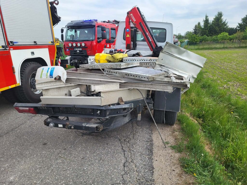 Uszkodzony ciągnik z przyczepą załadowaną materiałami budowlanymi stoi na poboczu drogi, obok wozu strażackiego.