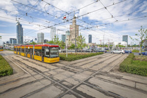 Tramwaj w kolorze żółto-czerwonym przejeżdża przez skrzyżowanie w miejskim otoczeniu z nowoczesnymi wieżowcami w tle.