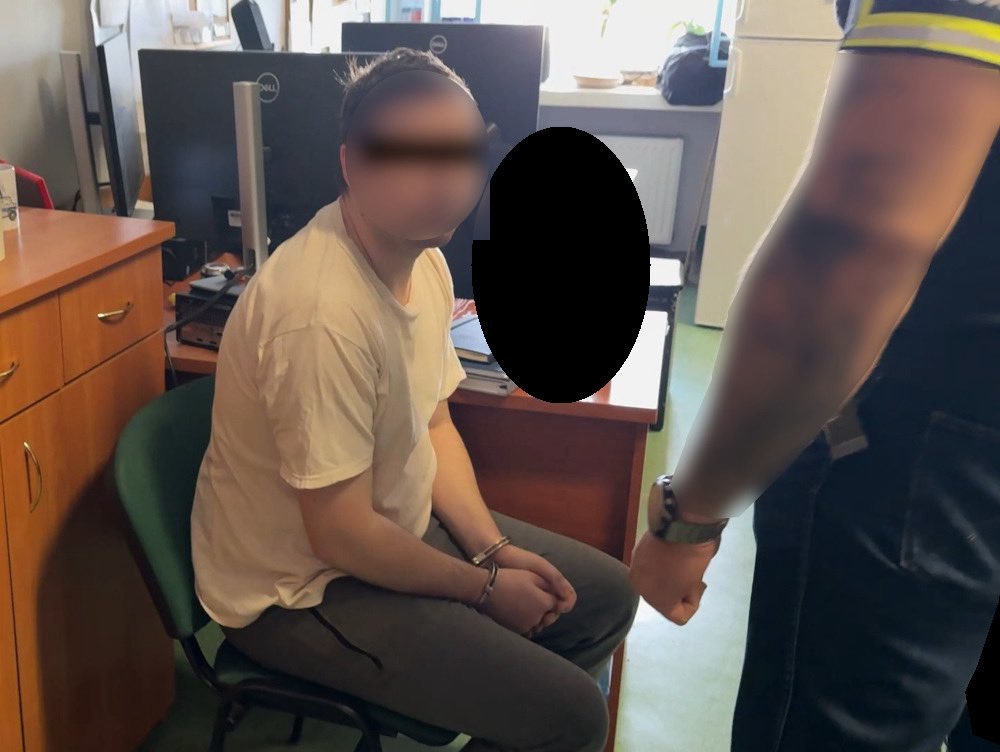 Młody mężczyzna w białej koszulce siedzi na krześle przy biurku, z rękami skuty w kajdankach. Policjant stoi obok.