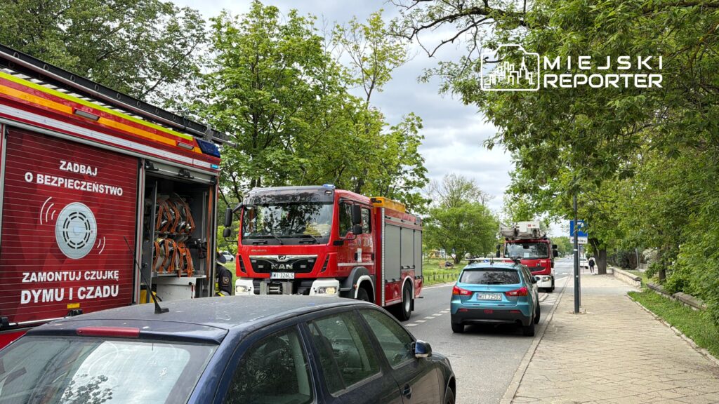 Czerwony wóz strażacki zatrzymany na ulicy, z otwartą klapą i widocznym sprzętem, w otoczeniu zieleni.